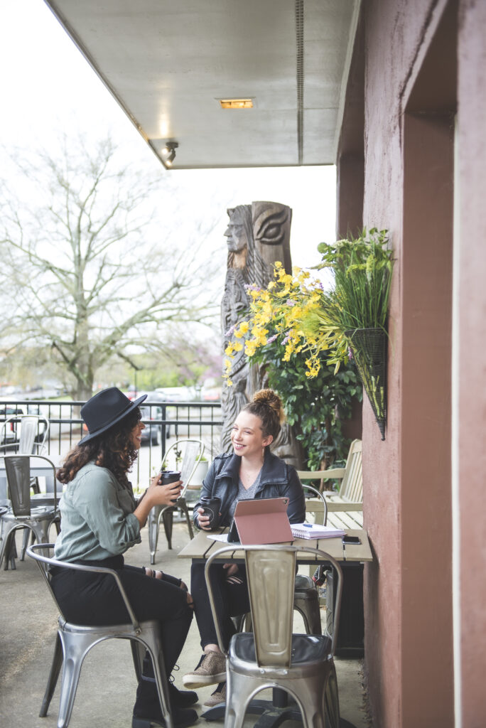 Two women sitting outside a coffee shop chatting over drinks with a laptop and notebooks on the table, yellow flowers and patio seating in the background.
