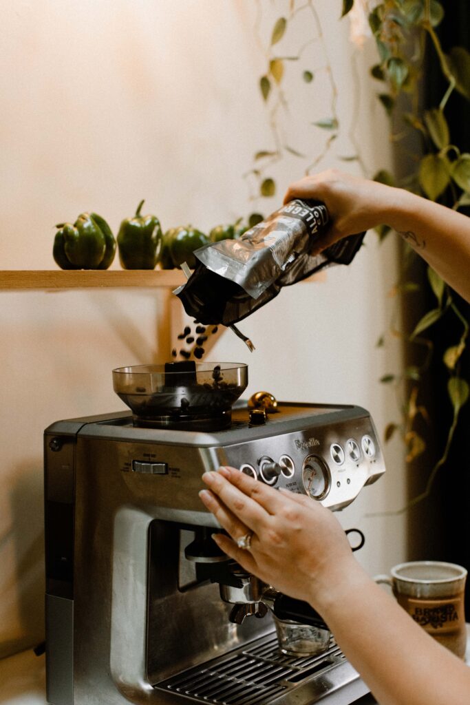 Hand pouring coffee beans into a Breville espresso machine with green bell peppers and trailing plants in the background.
